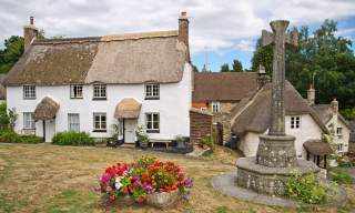 A quaint rural scene with white, thatched-roof cottages surrounded by greenery. A stone cross and colourful flower bed add charm under a partly cloudy sky.