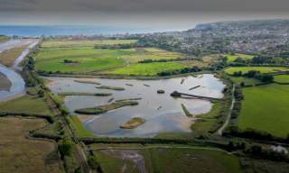 Aerial view of a scenic wetland area with ponds, surrounded by vibrant green fields and distant hills. A tranquil landscape under a clear sky.