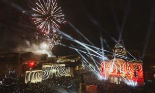 An overview of Hull City Hall and the Maritime Museum, lit up at night with spirals and flashlights, whilst fireworks explode overhead. A large crowd is gathered to watch the display.