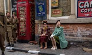 Actors dressed in 1940s clothing sit beside a vintage British red telephone box on a cobbled street, surrounded by period military uniforms and retro advertising signs.