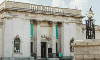 Front exterior of the Ferens Art Gallery in Hull, showing its neoclassical stone façade with tall columns, decorative carvings and entrance banners on a bright day, with the nearby historic lamp feature in the foreground.