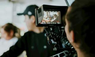 A man looks through a filming camera in the foreground as the people being filmed are blurred in the background.