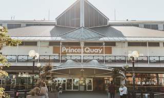 Exterior view of Princes Quay shopping centre in Hull, showing the main entrance, large glass roof structure, and outdoor walkway with trees and decorative lighting.