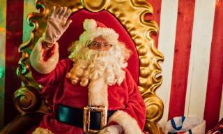 A jolly looking Santa waves as he sits on a plush throne in his grotto