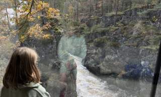 Et barn sitter ved et stort vindu og ser ut på Kvåsfossen i Lyngdal. Elva fosser ned gjennom kløften omgitt av høstfargede trær.