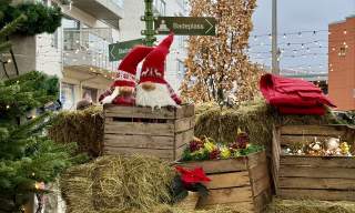 En juledekorert scene i sentrum av Lyngdal, med halmballer, trekasser fylt med julepynt, to nisser, røde julestjerner og et grønt veiviserskilt som peker til lokale attraksjoner. Små lyslenker og et tre i bakgrunnen gir førjulsstemning.