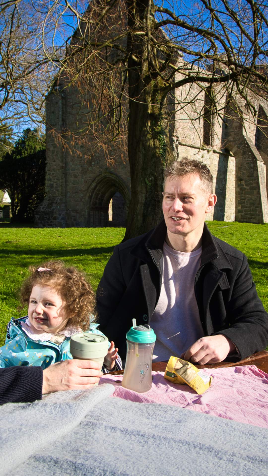 Family enjoying a picnic on a sunny day at a picnic table in front of Grey Abbey, County Down.