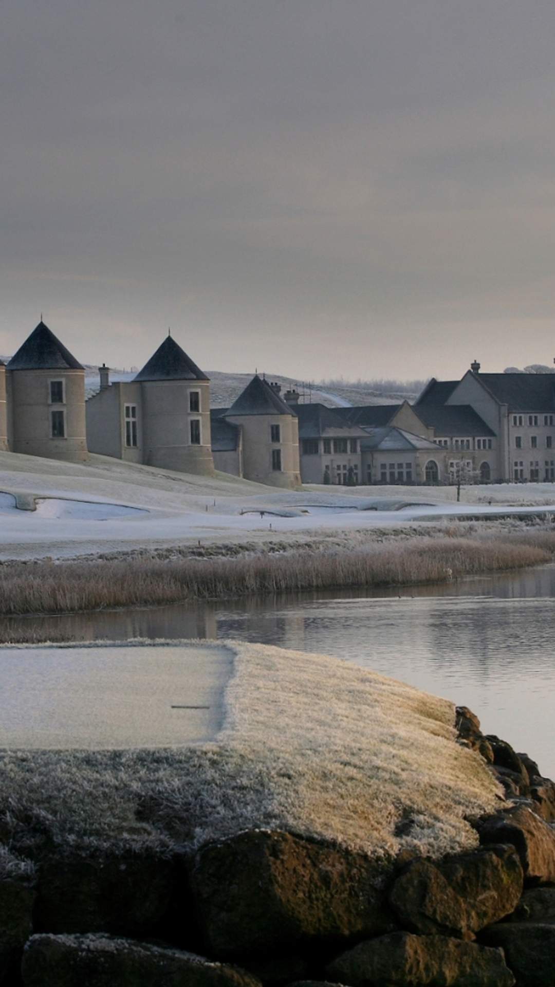 Snow-covered Lough Erne Resort reflected in a calm lake at winter sunrise, with two swans gliding across the water.