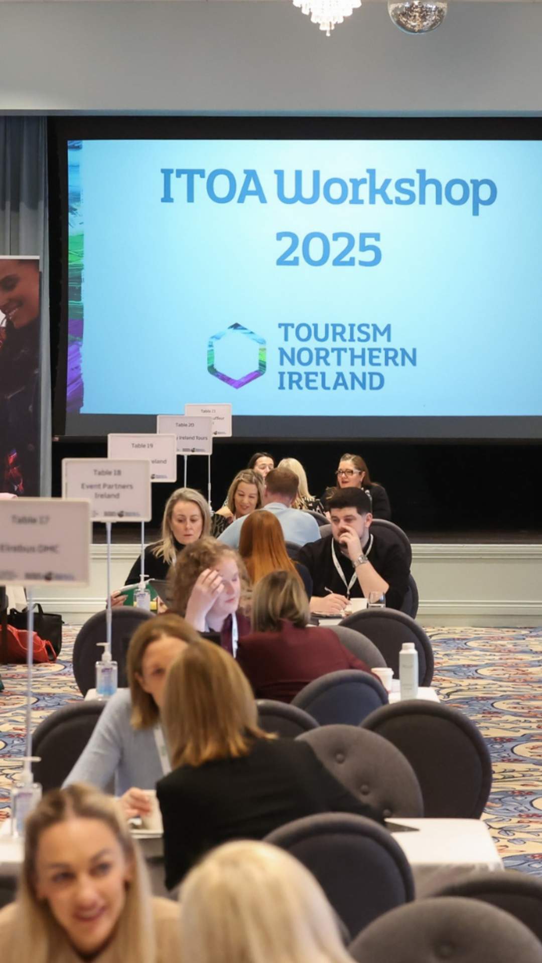 Delegates take part in one-to-one business meetings at the ITOA Workshop 2025, seated at numbered tables in a conference venue. A large screen displaying “ITOA Workshop 2025” and Tourism Northern Ireland branding is visible at the front of the room, with exhibition banners and decorated Christmas trees on either side.