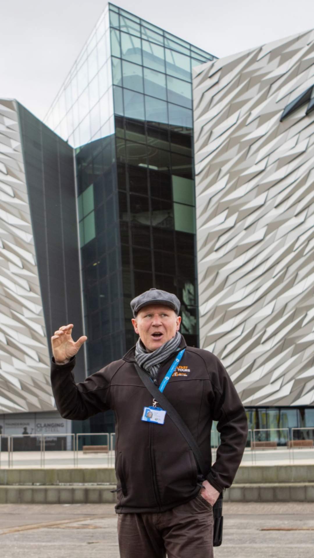 A tour guide speaks to a small group of visitors in front of the iconic Titanic Belfast building, with its distinctive angular silver façade and glass atrium visible in the background at Titanic Quarter, Belfast.