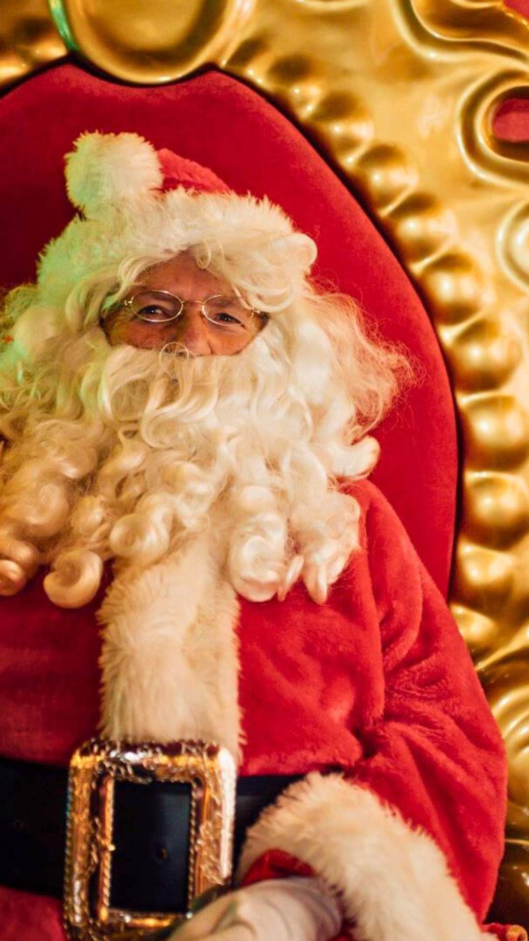 A jolly looking Santa waves as he sits on a plush throne in his grotto