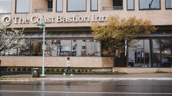 A person walks past The Coast Bastion Inn on a rainy day, with wet pavement and trees lining the street.