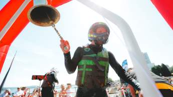 A racer wearing a helmet and life jacket rings a finish bell beneath a red arch as a crowd cheers along the waterfront behind him.