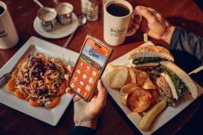 Two people sitting at a table with two plates of food and cups of coffee. One person is holding a phone with the Galena Country app showing.