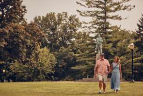 Couple walking in a park with a statue and trees behind them.