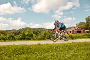 Two people riding bicycles on a road with a red barn behind them.