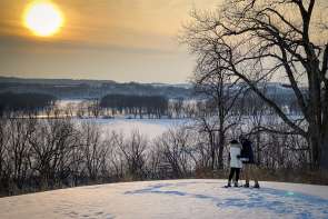 Two people in winter, looking over a bluff on a snowy hill. The sun is setting in the distance.