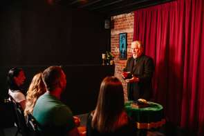 A magician performing on stage with a red curtain behind him, in front of an audience of people.