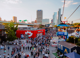 A large crowd moves through the bustling Summerfest grounds in Milwaukee during the day, with the downtown skyline visible in the background. A red building with the Summerfest smiley-face logo serves as the festival store. Above, colorful gondolas on the Skyglider transport people across the venue. The scene is filled with booths, food stands, and signage under a clear sky.