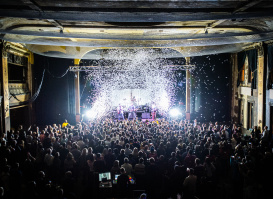 Interior view of Turner Hall ballroom during a live concert, showing a packed crowd of hundreds of people facing an illuminated stage. The historic venue's ornate architecture is visible with decorative columns, balconies, and detailed ceiling work. Dramatic stage lighting creates bright white beams cutting through the dark space, with confetti or snow effects falling from above.