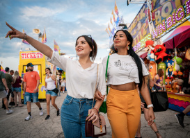 Two young women walk arm-in-arm through the Wisconsin State Fair, with one pointing excitedly ahead. Carnival rides, game booths, and crowds create a festive atmosphere in the background.