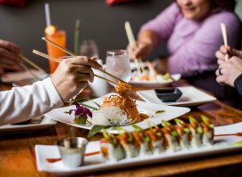 Diners gather around a table sharing sushi and Japanese-inspired dishes, using chopsticks to sample fresh rolls and plated seafood. The restaurant setting creates a warm, social atmosphere centered on food and conversation.