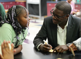 A man in a dark suit and glasses sits at a table, smiling and talking with a young girl who is leaning in to listen. They appear to be engaged in a learning activity, with papers or small items on the table, while other children and classroom materials are visible in the background.