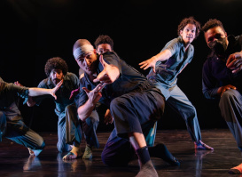 A group of six dancers strike expressive, dynamic poses on stage under dramatic lighting, wearing modern, casual performance outfits.