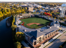 An aerial view of ABC Supply Stadium beside a winding river, surrounded by colorful autumn foliage and the city skyline. The baseball field sits at the center of the scene, with nearby streets, buildings, and a water tower visible under clear skies.