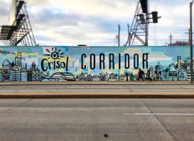 A large outdoor mural reading “Crisol Corridor” with illustrated cityscape elements in shades of blue and yellow. Two pedestrians walk past on the sidewalk in front of the mural under an overcast sky.