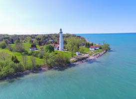 Aerial view of a white lighthouse and keeper’s buildings on a green lakeside point, surrounded by trees and lawns along the shoreline of Lake Michigan. Clear blue water stretches into the distance beneath a bright sky, with the lighthouse standing prominently above the landscape.