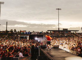 A performer in a red hoodie stands on an outdoor stage facing a massive crowd at sunset. Thousands of fans pack the venue, many holding up phones to capture the moment, while bright stadium lights and food stand signs glow in the background.