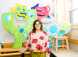 Hattie Grimm stands smiling beside colorful, oversized art pieces in a bright studio or gallery space. The whimsical sculptures feature bold shapes, patterns, and vibrant colors, highlighting a creative and playful artistic environment.