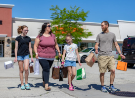 A family carrying shopping bags walks together through an outdoor shopping center on a bright, sunny day. The group smiles and talks as they cross the parking area, with storefronts, trees, and parked cars in the background.