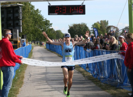 A runner raises their arms in celebration while crossing the finish line at a road race. Spectators line both sides of the course behind barriers, cheering as a race clock overhead displays the finishing time and volunteers hold the finish tape.