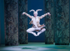 A dancer dressed as the White Rabbit leaps gracefully in midair, wearing a white costume with gold details and fluffy rabbit ears. The stage is softly lit in cool tones, with tall patterned columns in the background creating a whimsical, dreamlike setting.