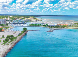 An aerial view of Reefpoint Marina along the Lake Michigan shoreline, featuring rows of docked boats, a protected harbor, and vibrant blue water. Waterfront buildings, green spaces, and a lakeside cityscape stretch beneath a sky filled with scattered clouds.