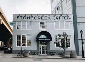 Exterior of Stone Creek Coffee’s white brick building with large front windows, black awning, and outdoor seating along the sidewalk.