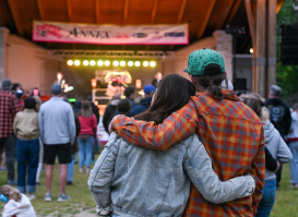 A couple stands arm in arm watching a live outdoor concert from the audience. Stage lights glow beneath a covered pavilion as community members gather on the lawn to enjoy the performance.