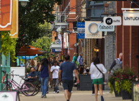 Pedestrians stroll past shops and outdoor dining areas along a bustling Historic Third Ward sidewalk, with teal streetlamps, storefront signs and string lights overhead.
