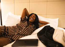 A person lying on a hotel bed, smiling and laughing with their arms resting behind their head. A closed laptop sits on the bed nearby, and a jacket is draped beside them. The room has soft lighting and a padded headboard, creating a relaxed, comfortable atmosphere.