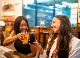 Two women smiling and clinking glasses of Sprecher soda at a wooden table inside a brewery
