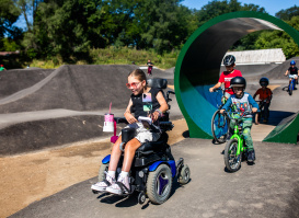 A smiling child using a power wheelchair rides along a pump track at a park, alongside other children on bicycles, with green play structures and trees in the background.