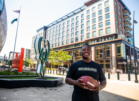 LeRoy Butler smiles while wearing a black ‘Milwaukee’ T-shirt holds a football in an outdoor plaza, with The Trade Hotel and a large deer sculpture visible behind him in Deer District.