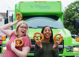 two friends standing in front of green tour bus showing off soft pretzels