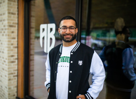 Nimesh "Nemo" Ruparel stands smiling outside a storefront wearing a black and white varsity-style jacket with "MILWAUKEE" text in green letters and "AB" logo. He has glasses, a beard, and is positioned in front of glass windows with "AB" lettering visible in the background. The setting appears to be an urban commercial area with brick architecture.