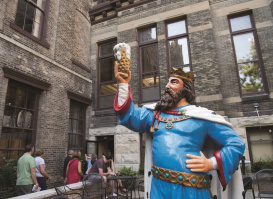 A colorful statue of King Gambrinus, the legendary beer king, holding up a frothy mug of beer in a celebratory pose outside the Historic Pabst Brewery in Milwaukee. People are gathered nearby in the brick courtyard.