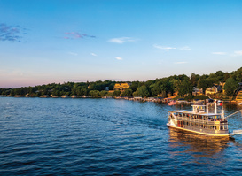 A tour boat cruises across a calm lake at sunset, with passengers aboard and shoreline homes nestled among trees in the background. Warm golden light reflects off the water, creating a scenic and relaxing lakeside view.