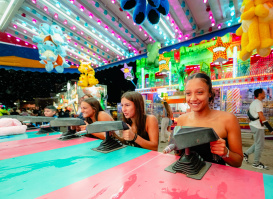 Three women smile while competing at a carnival squirt gun game beneath colorful lights and oversized stuffed animal prizes at the Wisconsin State Fair.