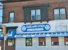 Front view of Oakland Gyros, a two-story brick building with a white-painted lower level and blue signage reading “Oakland Gyros – Best Gyros in Town.” The storefront features blue awnings advertising kabobs, lamb, spinach pie, and Athenian chicken